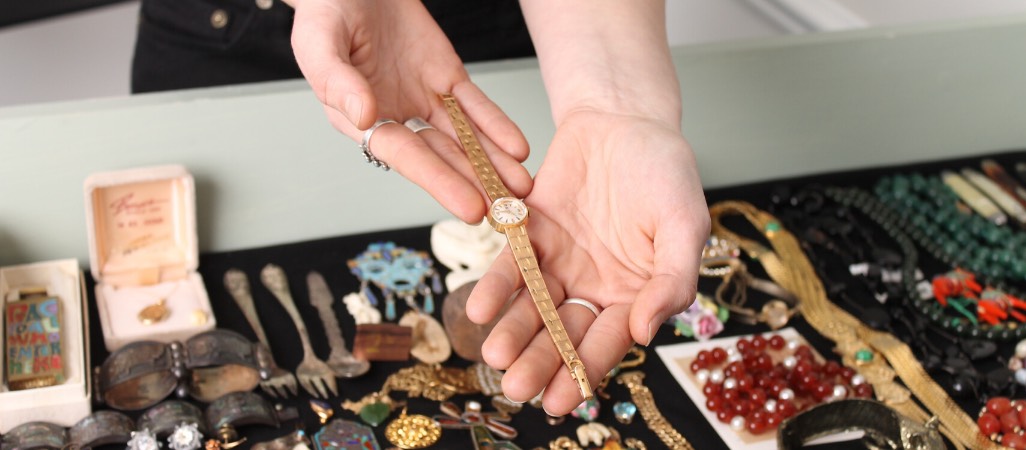 two hands holding up a gold woman's watch with other trinkets below