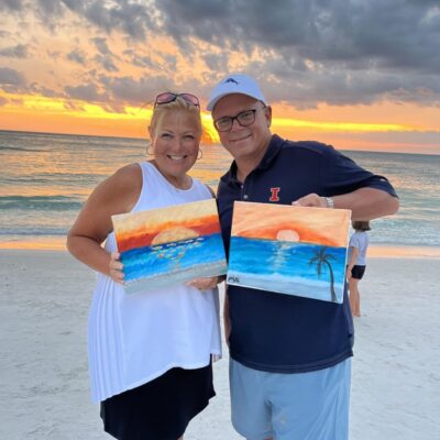 couple on beach holding up beach scene paintings