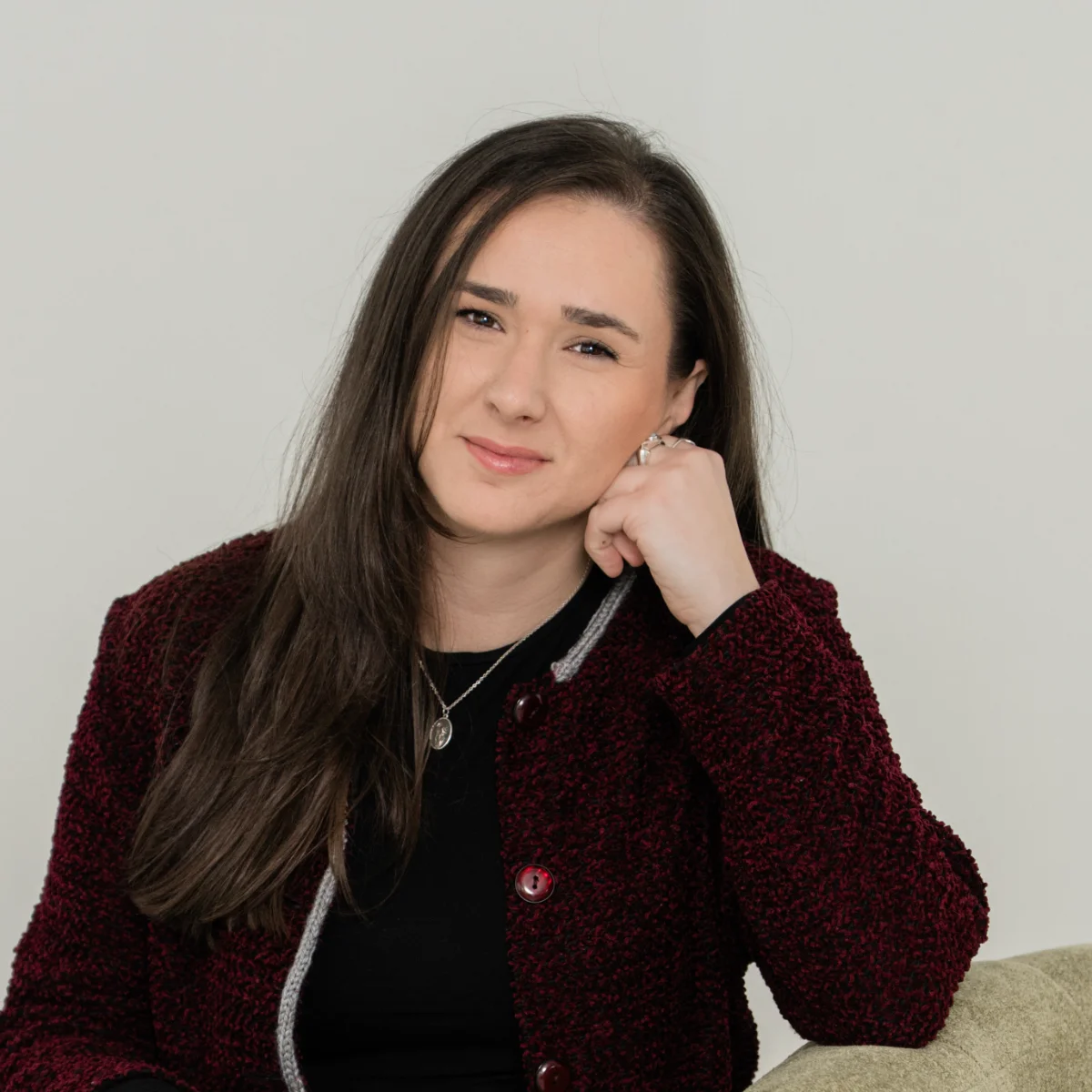 Ariel Regner, wearing a burgundy blazer, smiles warmly while posing with her cheek resting on her hand.