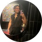 Alvaro Lozano, wearing a dark star-cutout top, smiles brightly while taking a mirror selfie in a stylish, dark interior with colorful abstract art visible behind him.